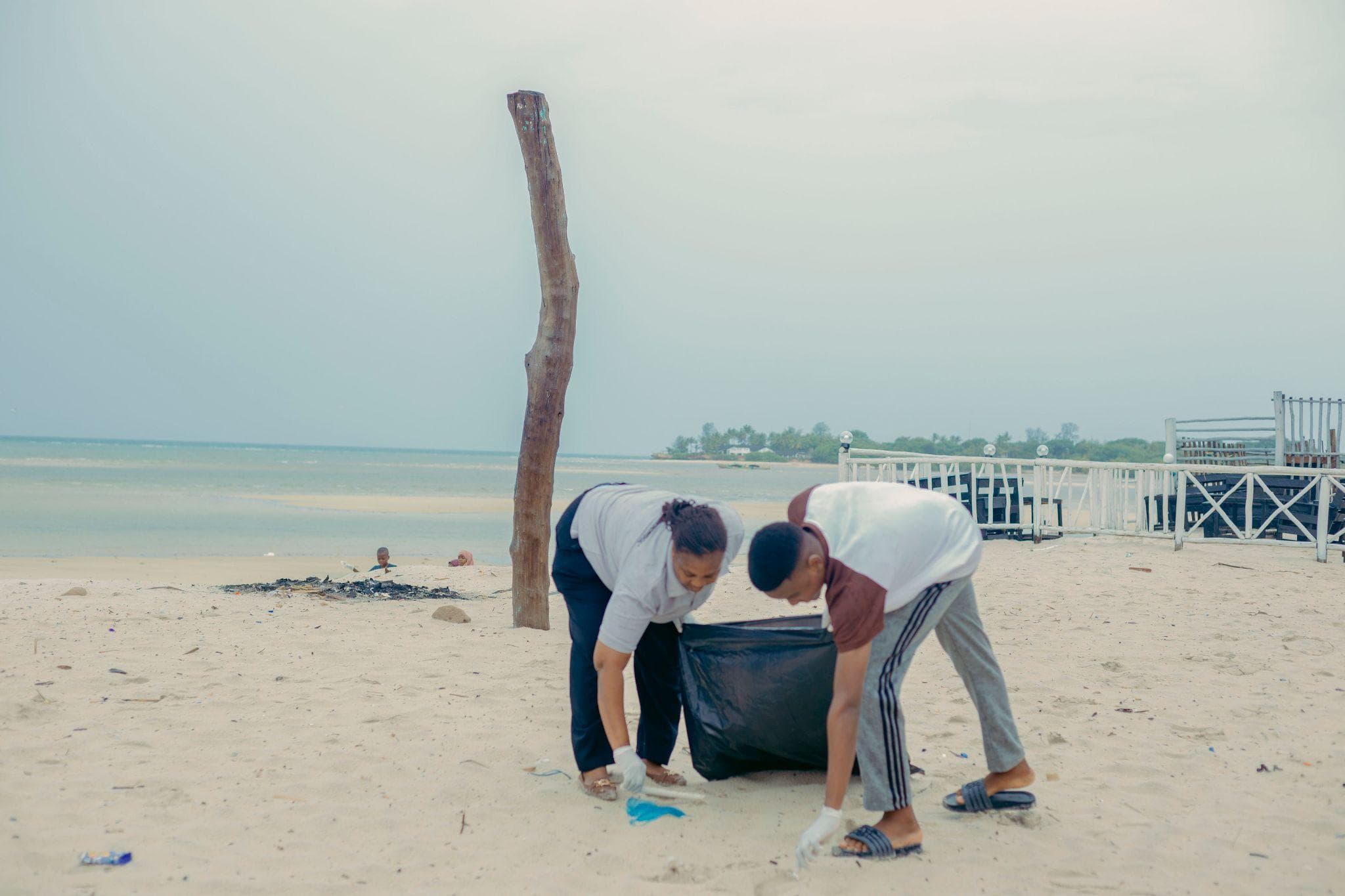 Two volunteers collecting litter into a large bag on the shoreline.